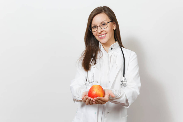 Smiling confident beautiful young doctor woman with stethoscope, glasses isolated on white background. Female doctor in medical gown holding red apple. Healthcare personnel, health, medicine concept. - Fotó, kép