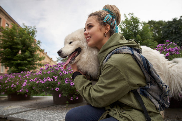 Cute young teen girl with dreadlocks on her head in a light green jacket and a backpack walks through a blooming Park with her four-legged friend Samoyed dog. - Фото, изображение