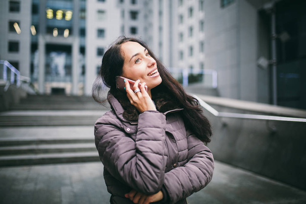 young girl talking on mobile phone in courtyard business center. girl with long dark hair dressed in winter jacket in cold weather speaks on phone on background buildings made of glass and concrete - Zdjęcie, obraz