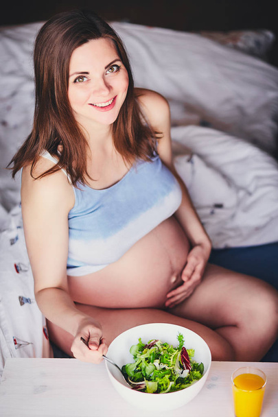 Pregnant smiling girl in T-shirt sit on bed and eat salad with green and red leaves, drink orange, pumpkin and carrot juice. Young woman keep diet during pregnancy. Healthy food for expectant mothers. - Photo, Image