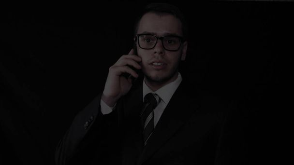 Handsome young businessman wearing glasses, talking on the phone in a suit, black background. - Photo, image