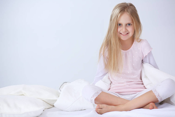 Little girl in the bedroom is sitting on the bed. Little girl is wearing a pajamas and sitting in bed - Fotografie, Obrázek