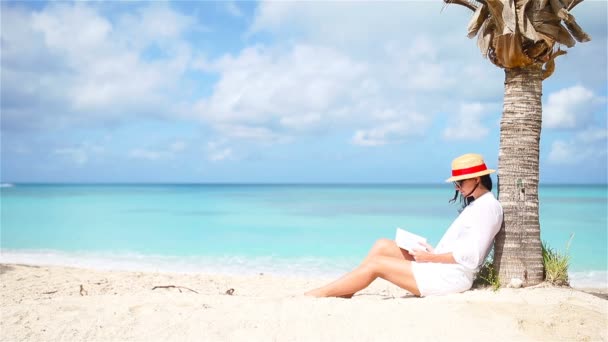 Jovem mulher lendo livro sob palmeira na praia
 - Filmagem, Vídeo