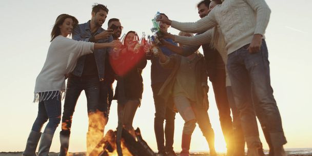 Young Friends Making A Toast With Beer and having fun Around Campfire at beach party on autumn day - 写真・画像