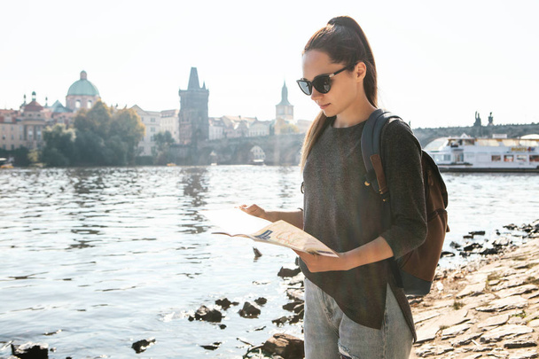 A young beautiful girl stands and looks at the map next to the Vltava River with the amazing old architecture of Prague in the background with a sunny spring day - Photo, image