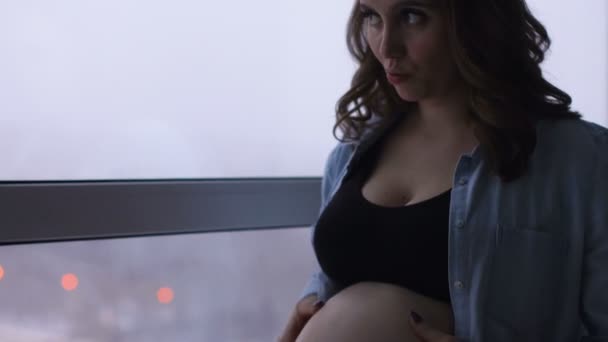 Yüksek bir irtifada bulunan büyük bir panoramik pencere, mutlu bir hamile kadın durumda. Ellerini karnına dokunur ve gülümseyerek karnında bebek ile iletişim kurar. Porteut - Video, Çekim