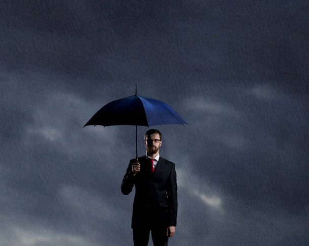 Businessman with umbrella standing over stormy background. Business, protection, crisis concept.   - Photo, image