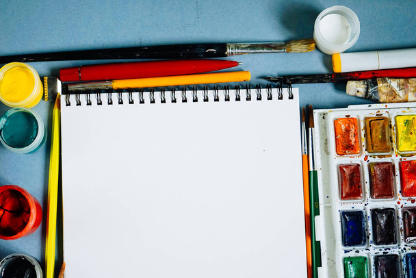 on a blue background there are paints, brushes, pencils and a sheet of white paper - Photo, Image