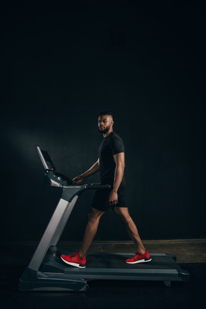 молодой африканский американский спортсмен, тренирующийся на беговой дорожке и смотрящий в камеру на черном
 - Фото, изображение