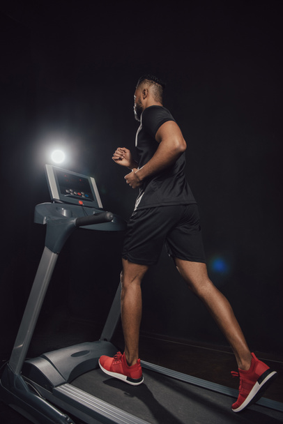 lage hoekmening van jonge Afro-Amerikaanse sportman joggen op een loopband op zwart - Foto, afbeelding