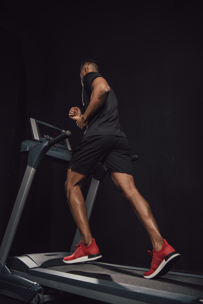 low angle view of young african american sportsman running on treadmill isolated on black - 写真・画像