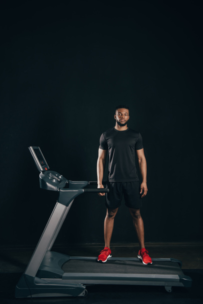 junger afrikanisch-amerikanischer Sportler steht auf dem Laufband und schaut in die Kamera auf schwarz - Foto, Bild