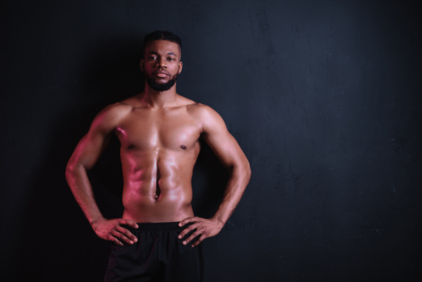 muskulöser Mann mit nacktem Oberkörper, der mit den Händen auf der Taille steht und isoliert auf schwarz in die Kamera schaut - Foto, Bild
