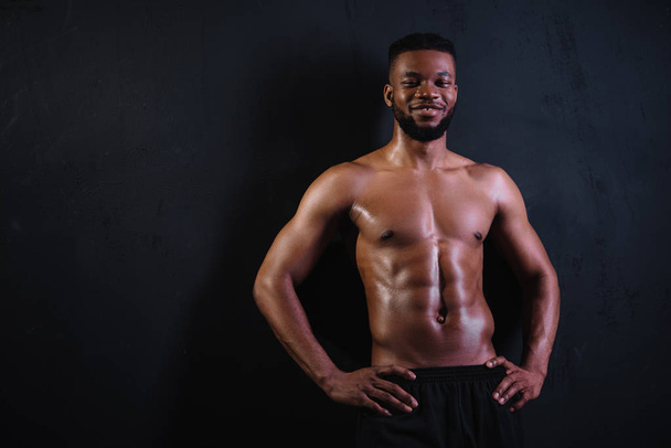 hemdloser muskulöser junger afrikanisch-amerikanischer Mann, der mit den Händen auf der Taille steht und auf schwarz in die Kamera lächelt    - Foto, Bild