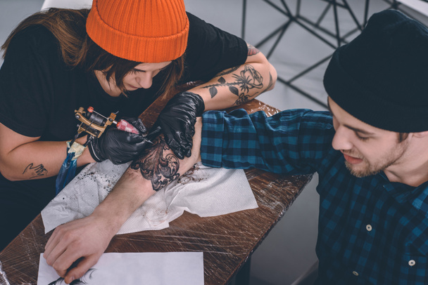 Hombre joven y maestro del tatuaje femenino durante el proceso de tatuaje en el estudio
 - Foto, Imagen