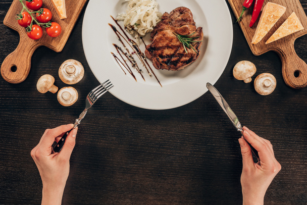 oříznutý obraz ženy držící vidličku a nůž nad deskou s hovězí steak - Fotografie, Obrázek
