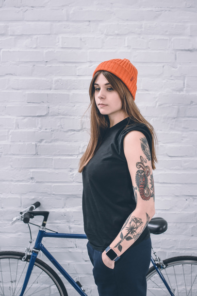 Chica con estilo con el brazo tatuado de pie junto a la bicicleta por la pared
 - Foto, Imagen