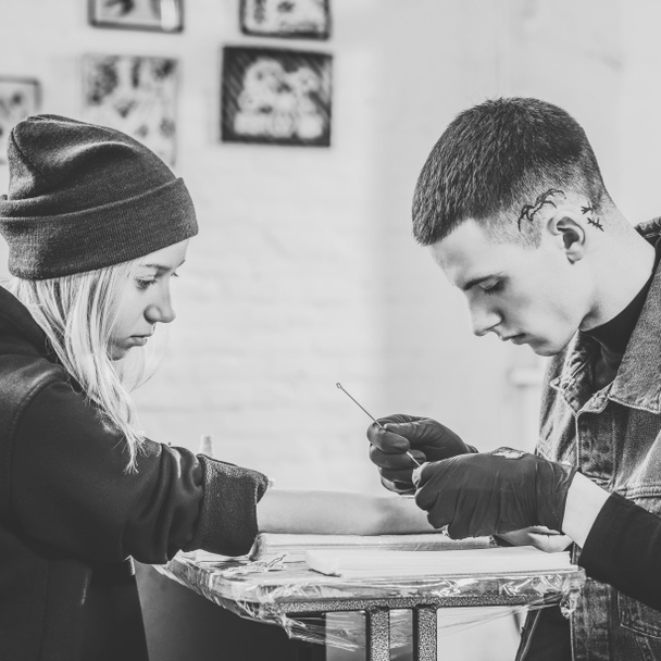 Black and white photo of tattoo artist in gloves working on female arm piece in studio - Photo, Image