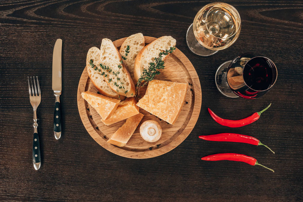 top view of parmesan cheese with baguette slices on wooden board, chili peppers and wine on table - Photo, image