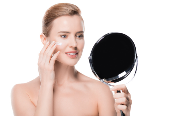Женщина наносит крем на лицо, глядя на зеркало, изолированное на белом
 - Фото, изображение