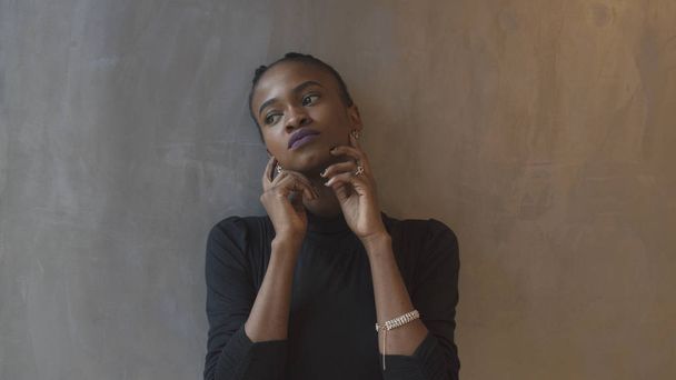 Attractive calm african woman in black dress touching her face and posing while looking aside and leaning on the gray wall. - Fotografie, Obrázek