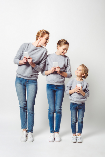 smiling mother and daughters in similar clothing with smartphones isolated on grey - Foto, afbeelding