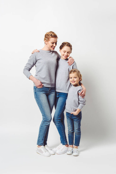 mother and daughters of different generations in similar clothing hugging each other isolated on grey - Фото, изображение