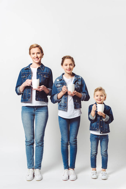lachende moeder en dochters in soortgelijke denim kleding met cups van warme dranken geïsoleerd op grijs - Foto, afbeelding