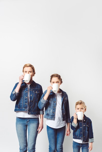 portrait of mother and daughters in similar denim clothing drinking hot drinks isolated on grey - Fotografie, Obrázek
