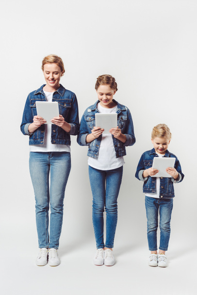 Lächelnde Mutter und Töchter in ähnlicher Jeanskleidung, die in einer Reihe stehen und Tabletten isoliert auf grau verwenden - Foto, Bild