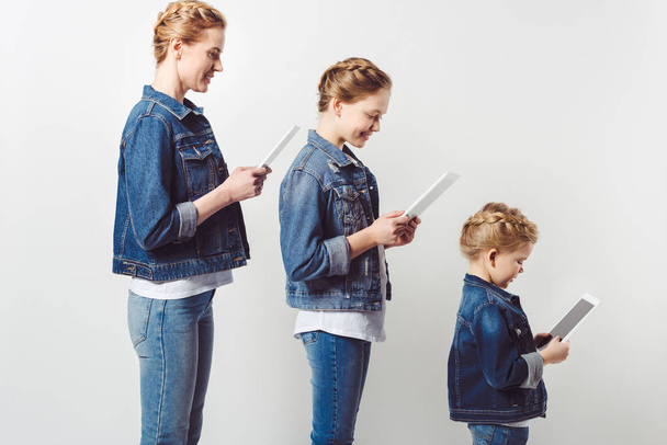 mother and daughters in similar denim clothing standing in row and using tablets isolated on grey - Foto, Imagem