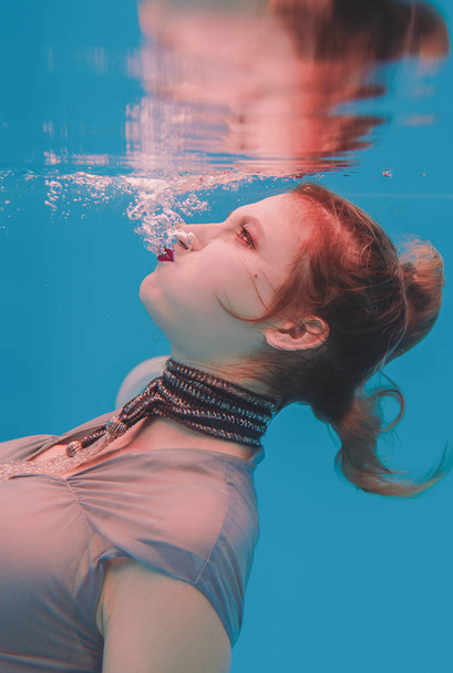 Amazing beautiful art surreal portrait of young angry woman in grey dress and beaded scarf underwater in the swimming pool - Fotoğraf, Görsel