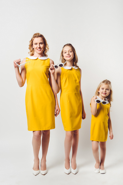 happy mother and daughters in similar retro style yellow dresses looking at camera on white - Фото, изображение