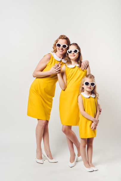 mother and daughters in similar retro style yellow dresses and sunglasses on white - Foto, afbeelding