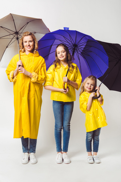 Szczęśliwa matka i córki w żółty płaszcze z parasolami, stojących w rzędzie na białym - Zdjęcie, obraz