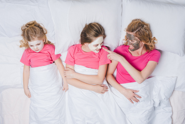 top view of happy mother and daughters lying in bed together with skincare masks  - Photo, Image