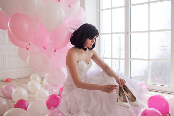 Young woman in wedding dress in luxury interior with a mass of pink and white balloons. Hold in hands her white shoes - Φωτογραφία, εικόνα