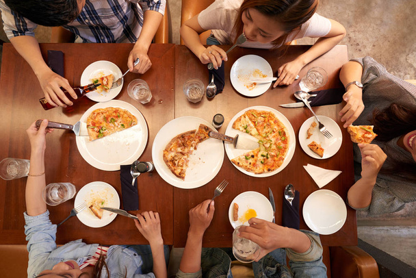 Grupo de jóvenes asiáticos comiendo pizza en una gran mesa en la cafetería, vista desde arriba
 - Foto, Imagen