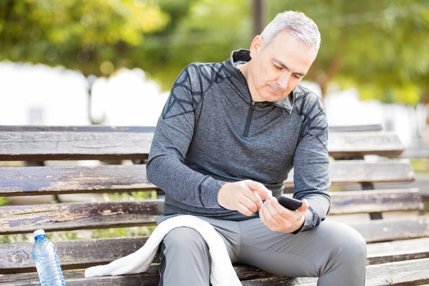 Hombre hispano maduro bien parecido sentado en un banco del parque y usando un teléfono inteligente después de hacer ejercicio
 - Foto, Imagen