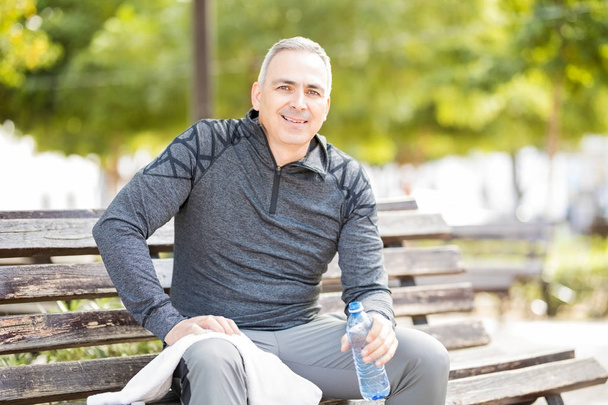 Good looking middle aged man sitting on a park bench with a water bottle after exercise session - Photo, Image