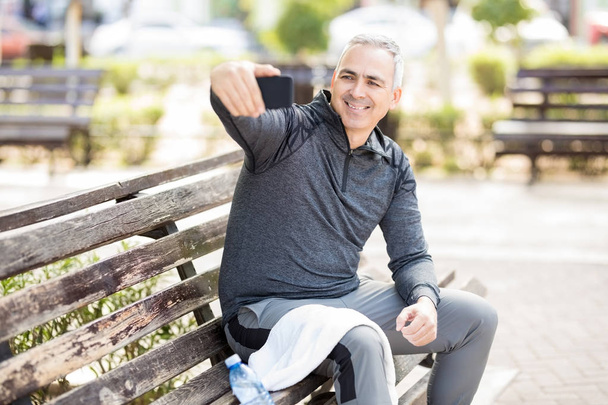 Smiling mature man sitting on park bench after jogging and taking a selfie with smartphone - Photo, Image