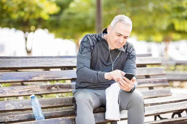 Feliz hombre de mediana edad sentado en el banco y escuchando música con teléfono móvil después de hacer ejercicio al aire libre en un parque
 - Foto, Imagen