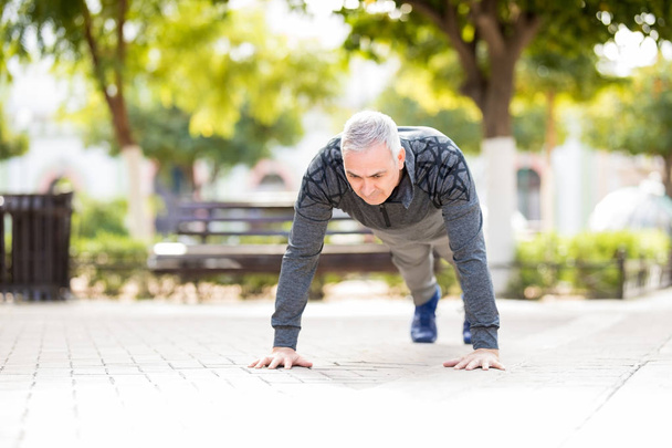 Starší muž s šedivými vlasy se zahřívá a dělá push up jako součást jeho výcviku v parku - Fotografie, Obrázek