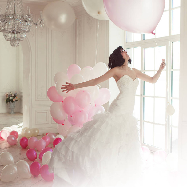 Young woman in wedding dress in luxury interior flies on pink and white balloons. - 写真・画像