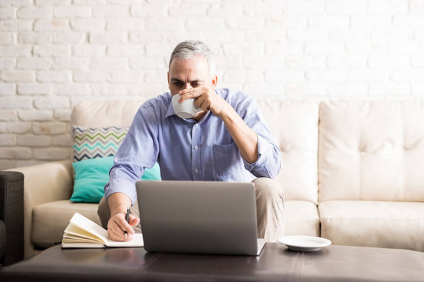 Hombre de mediana edad sentado en la sala de estar mirando a la computadora portátil tomando café y tomando notas en su diario
 - Foto, imagen