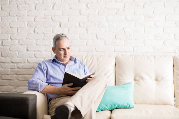 Good looking mature man relaxing on sofa and reading book at home interior - 写真・画像
