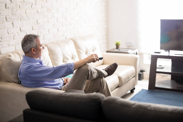 Mature man sitting in living room with remote control watching tv while relaxing at home - Foto, imagen