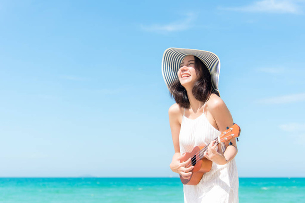 Summer Vacation. Smelling asian women relaxing and playing an ukulele on the beach, so happy and luxury in holiday summer, blue sky background. Travel and lifestyle Concept. - Photo, Image