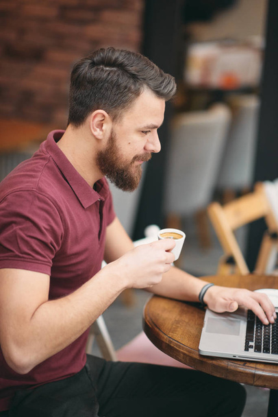 Człowiek pije kawę i za pomocą laptopa siedząc w kawiarni. - Zdjęcie, obraz