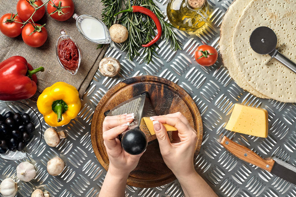 Las manos femeninas frotaron queso rallado en la pizza, ingredientes para cocinar pizza en la mesa de metal, vista superior
 - Foto, Imagen